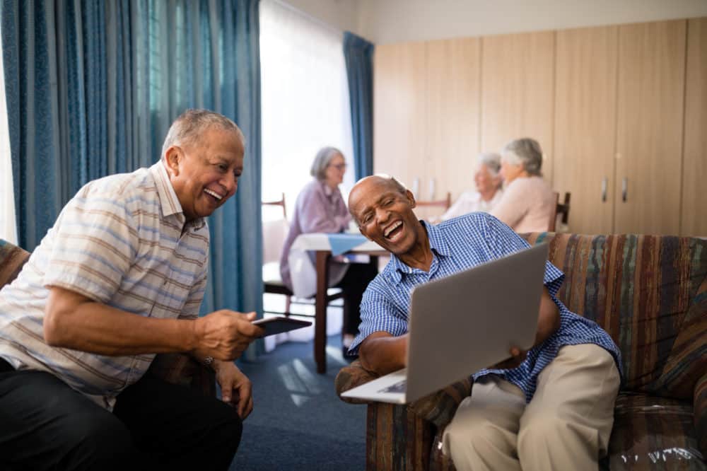 Happy senior friends looking at laptop while sitting on sofa against females in nursing home